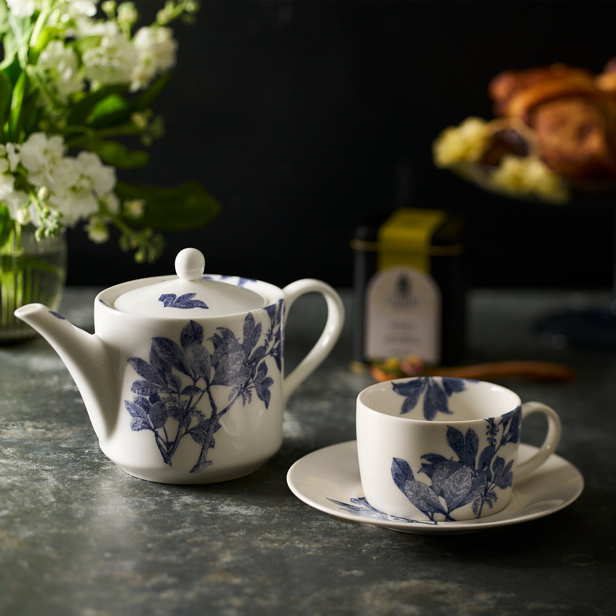A white porcelain teapot and Caskata Artisanal Home&#39;s Arbor Cups &amp; Saucers, Set of 2 with blue leaf designs, part of a stunning bone china collection, are on a countertop. A vase of flowers, a tin canister, and a plate with baked goods are in the background.