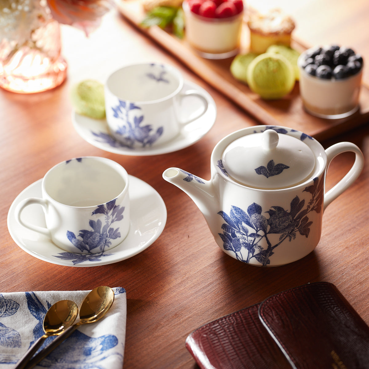 A wooden table with a tea set featuring a teapot and Caskata Artisanal Home Arbor Cups &amp; Saucers, Set of 2 with blue and white floral patterns, a napkin, two spoons, and a variety of desserts and fruits in the background. The elegant blue and white dinnerware complements the scene perfectly.