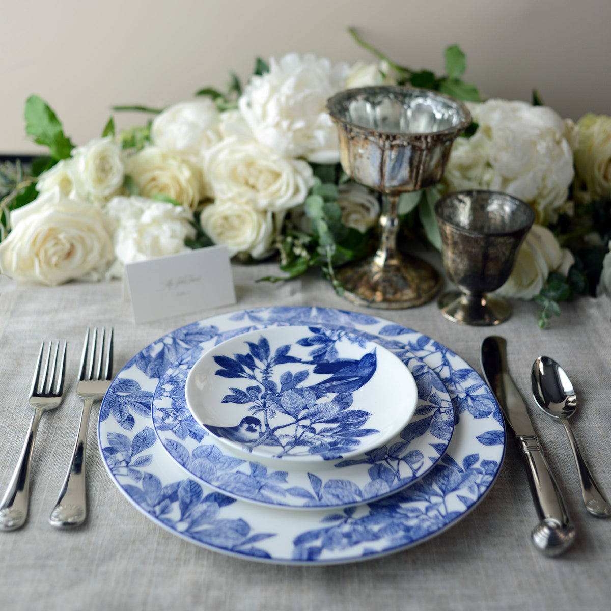 A table setting with blue and white floral-patterned plates, including a delicate Arbor Rimmed Salad Plate by Caskata Artisanal Home, silver cutlery, two ornate goblets, and a floral centerpiece of white roses and greenery. The premium porcelain dishes lend an heirloom feel to the scene. A place card is placed above the plates.