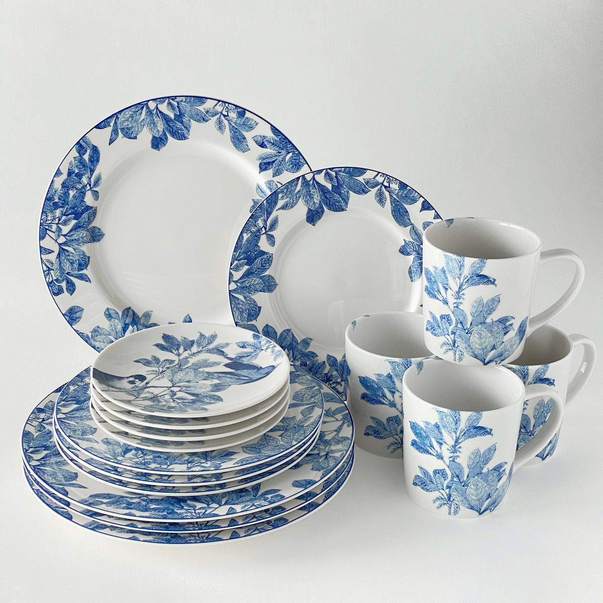 An Arbor Blue Birds Small Plates set from Caskata Artisanal Home, featuring heirloom-quality dinnerware with intricate botanical details, includes plates, bowls, and mugs arranged neatly against a white background.
