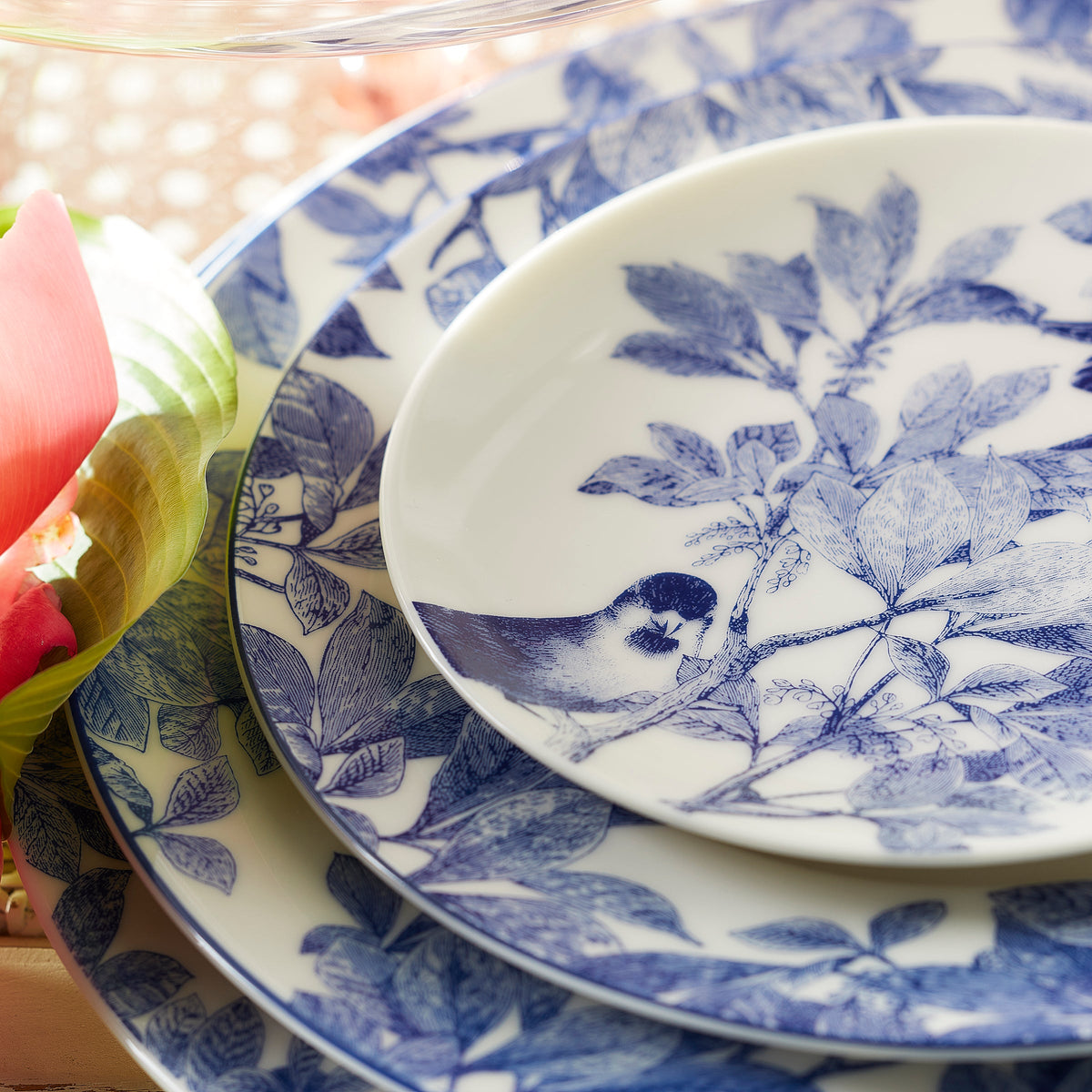 A set of white plates with blue floral and bird designs stacked on a table. The top plate, the Arbor Rimmed Charger Plate from Caskata Artisanal Home, crafted from bone china, features a bird perched on a branch among leaves. A pink flower and green foliage are partially visible nearby, showcasing intricate botanical details.