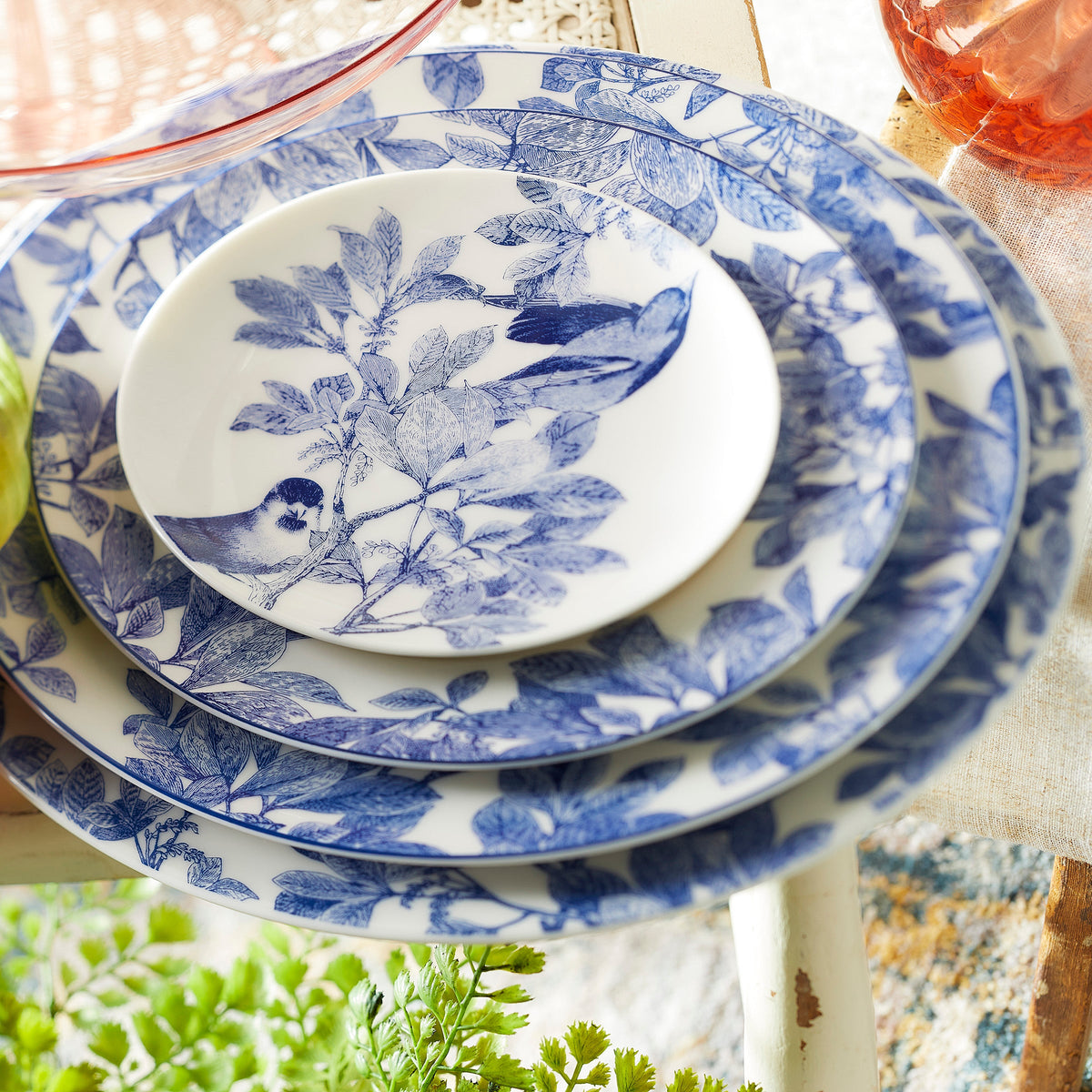 A stack of four blue and white bone china plates with intricate bird and foliage designs. The plates, showcasing exquisite botanical details, are arranged from largest to smallest. These are the Arbor Rimmed Charger Plates by Caskata Artisanal Home.
