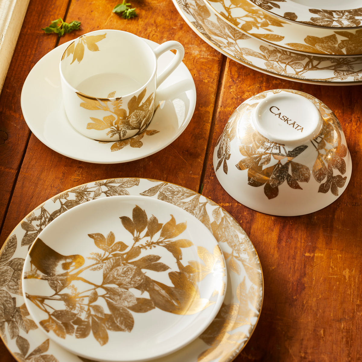 A collection of Arbor Gold dinnerware from Caskata, including a canapé plate on a salad plate, cup and saucer, small bowl and stack of plates on a rustic wooden tabletop.