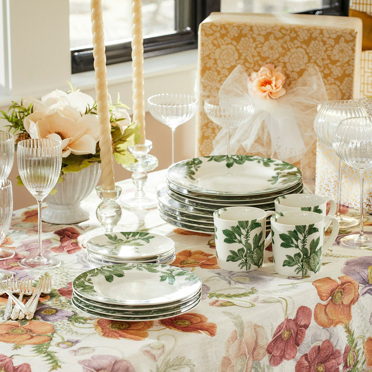 A botanical-themed table setting with Caskata Arbor Green Small Plates, matching mugs, crystal glassware, a floral centerpiece, candles, and a wrapped gift box with a bow.