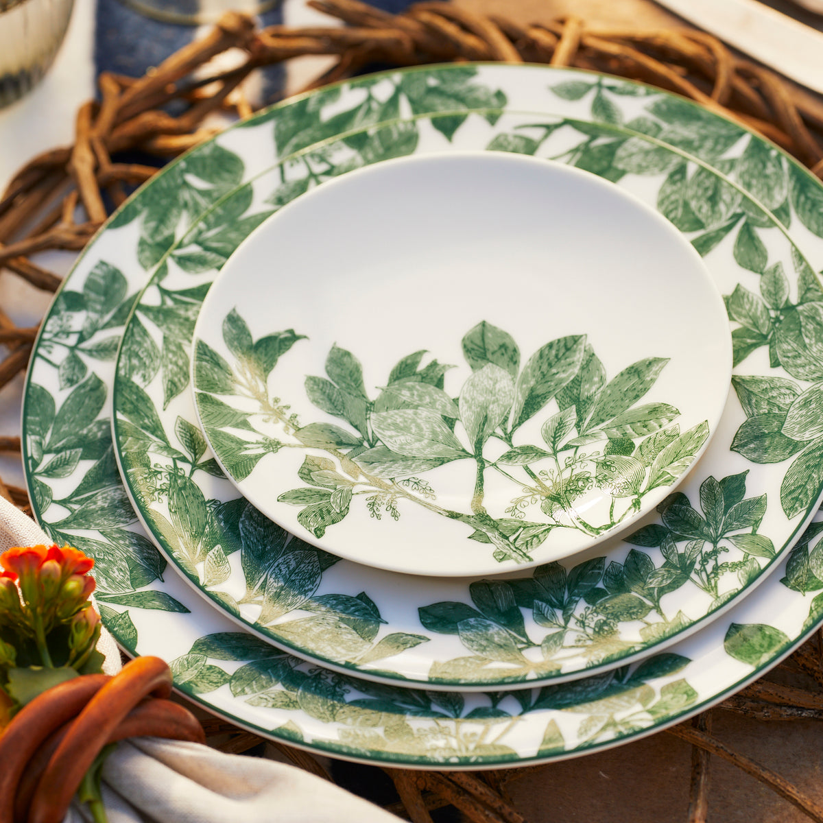 A set of three Caskata Arbor Green Small Plates with green leaf patterns is neatly stacked, placed on a braided charger. A small orange flower is partly visible in the foreground, adding a touch of color to the botanical dinnerware. These plates are also dishwasher safe for easy cleanup.