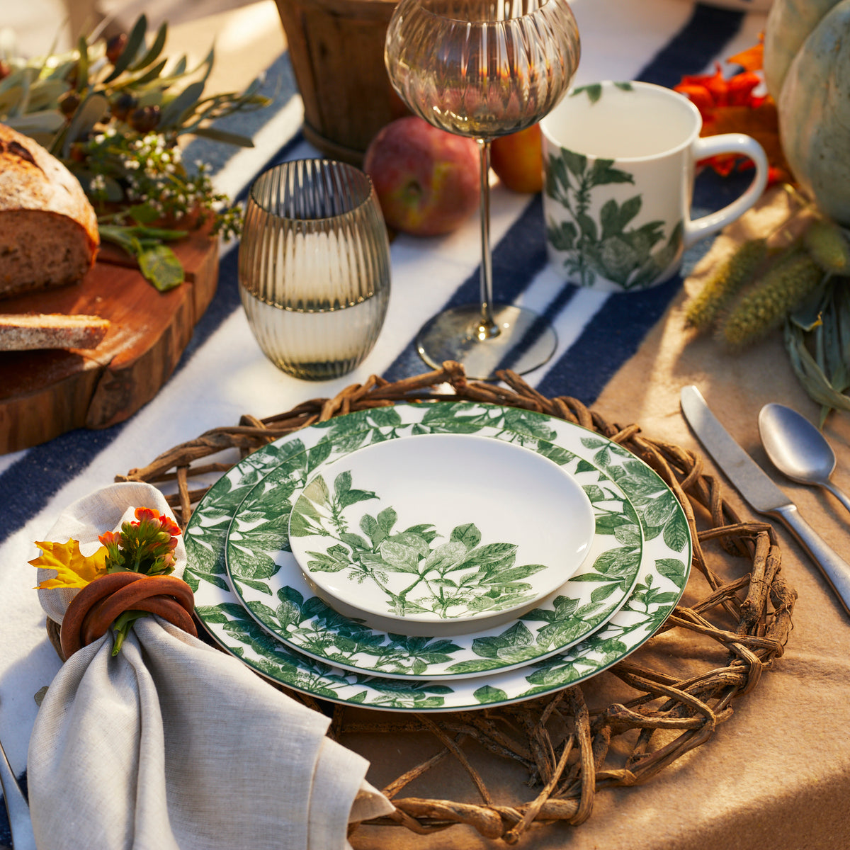 A table is set with green leafy patterned plates, a matching mug, glassware, and silverware on a woven placemat. Caskata Arbor Green Rimmed Salad Plates complement the setup. Bread, fruit, and decorative flowers are arranged nearby amidst an arbor of greenery.