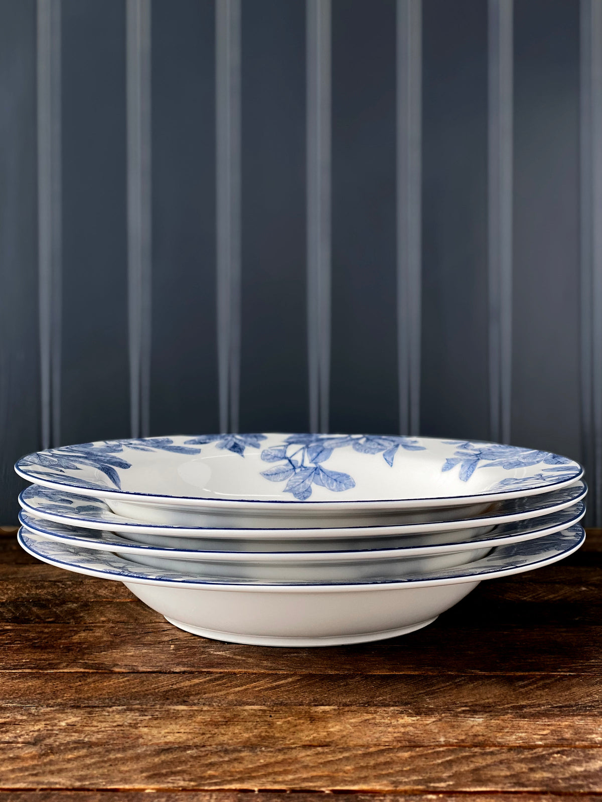Four classic Arbor Rimmed Soup Bowls by Caskata Artisanal Home, made of high-fire porcelain, with blue floral patterns, are neatly stacked on a wooden surface against vertical wooden panels. These white ceramic bowls are also dishwasher safe for your convenience.