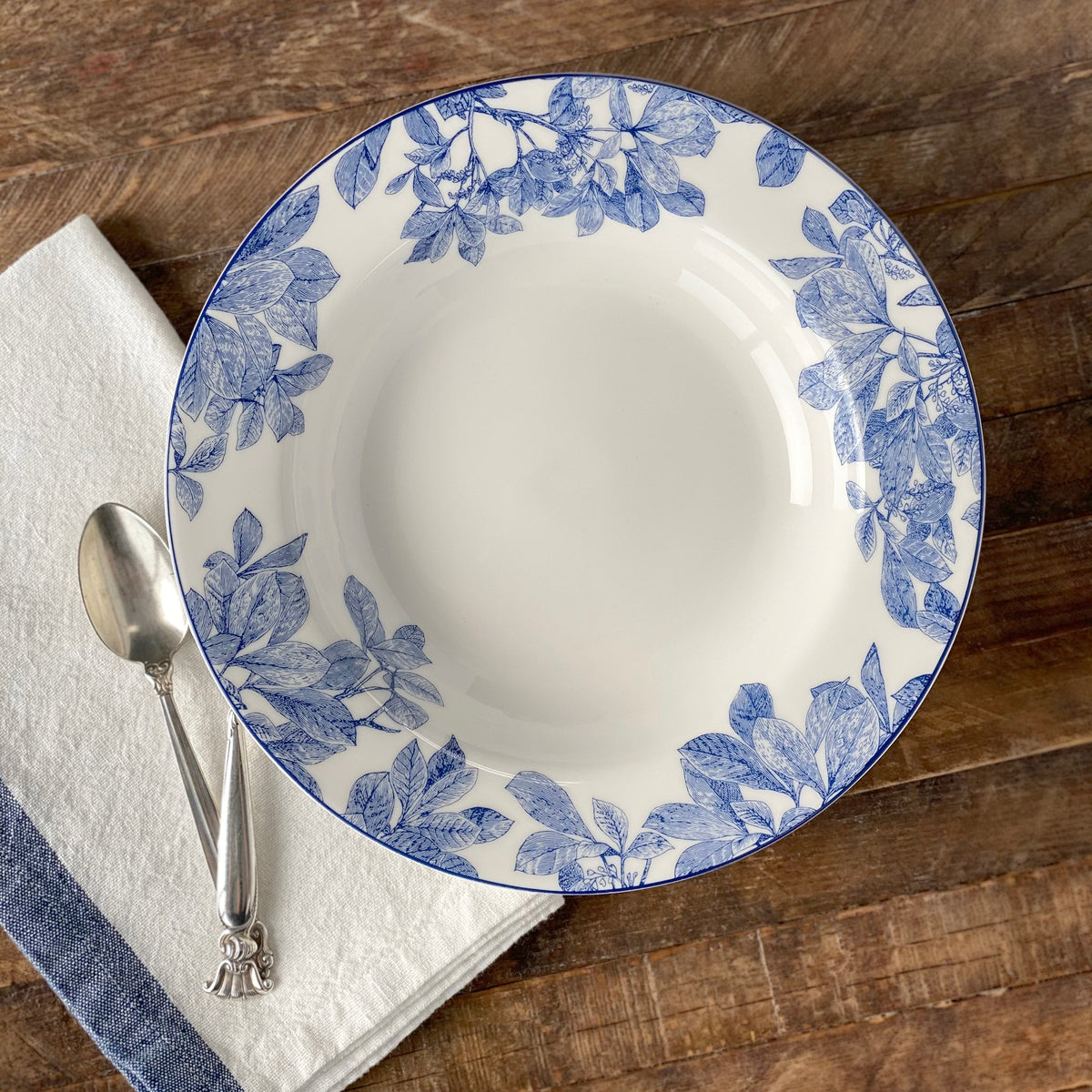 A classic **Arbor Rimmed Soup Bowl** made of high-fire porcelain with blue floral patterns by **Caskata Artisanal Home** is placed on a wooden table. To the left, there is a folded white cloth with a blue stripe, and a silver spoon rests on it. The bowl is not only elegant but also dishwasher safe.