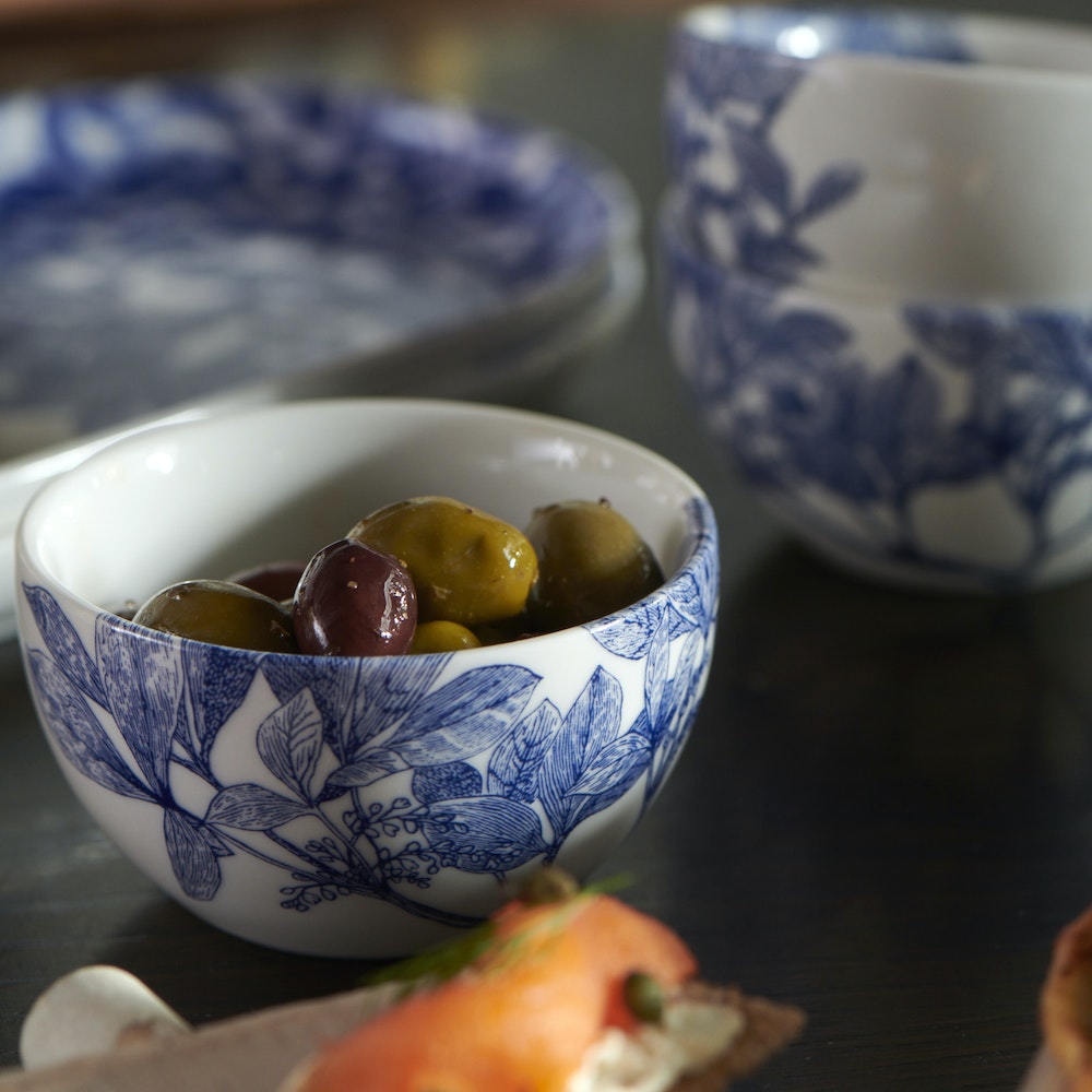A small Caskata Artisanal Home Arbor Snack Bowl with a blue floral pattern and leafy branches porcelain holds mixed olives. In the background, similar patterned bowls and plates are slightly out of focus.