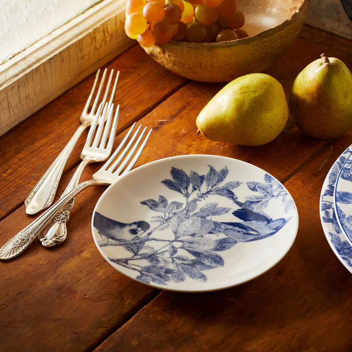 A white plate with botanical details and blue floral patterns, two pears, a bowl of grapes, and silver forks are arranged on a wooden table near a window. The Caskata Artisanal Home Arbor Blue Birds Small Plates enhance the rustic charm of the setting.