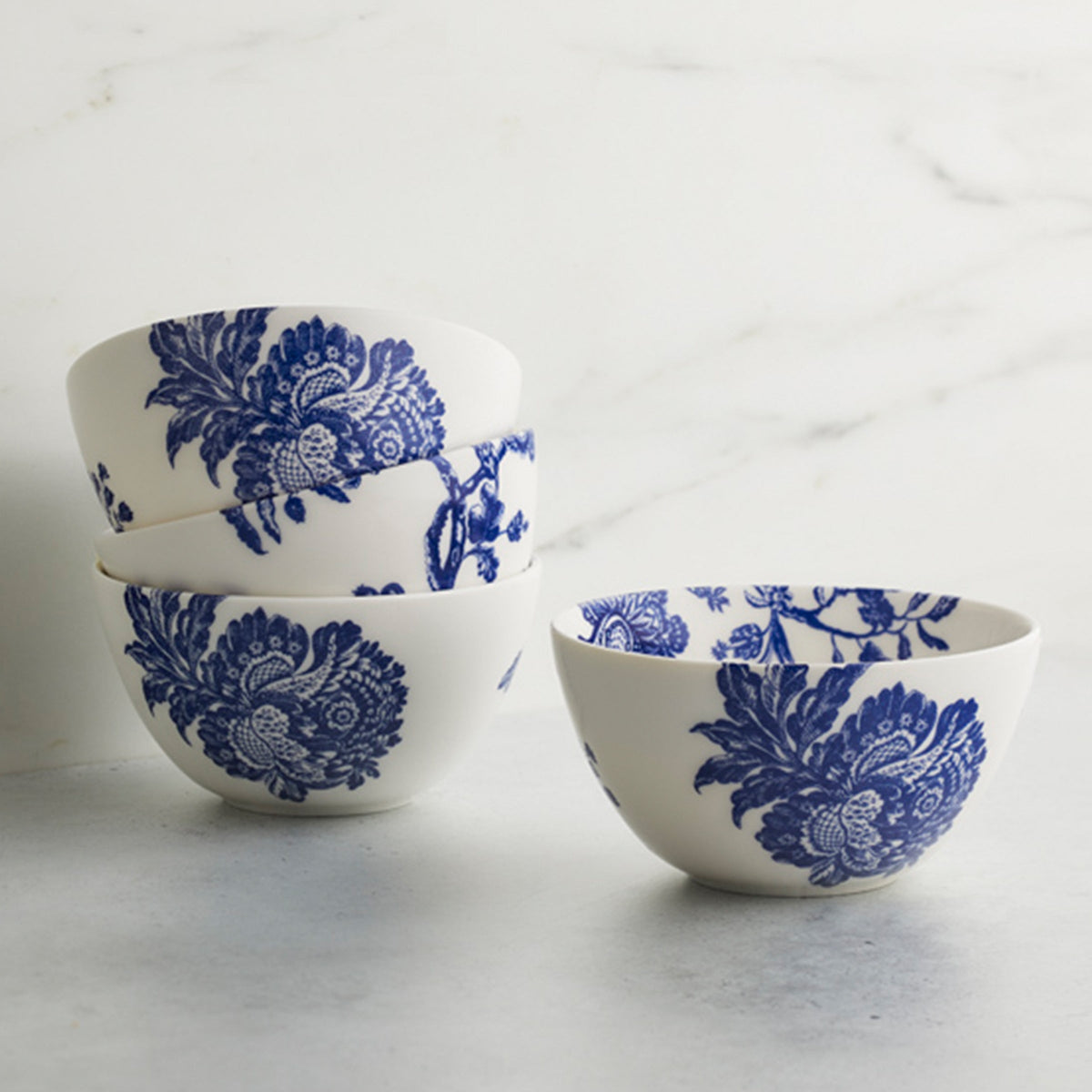 Four white Arcadia Caskata porcelain cereal bowls with blue floral patterns are stacked on a marble surface, evoking a sense of classic elegance inspired by the Williamsburg Foundation.