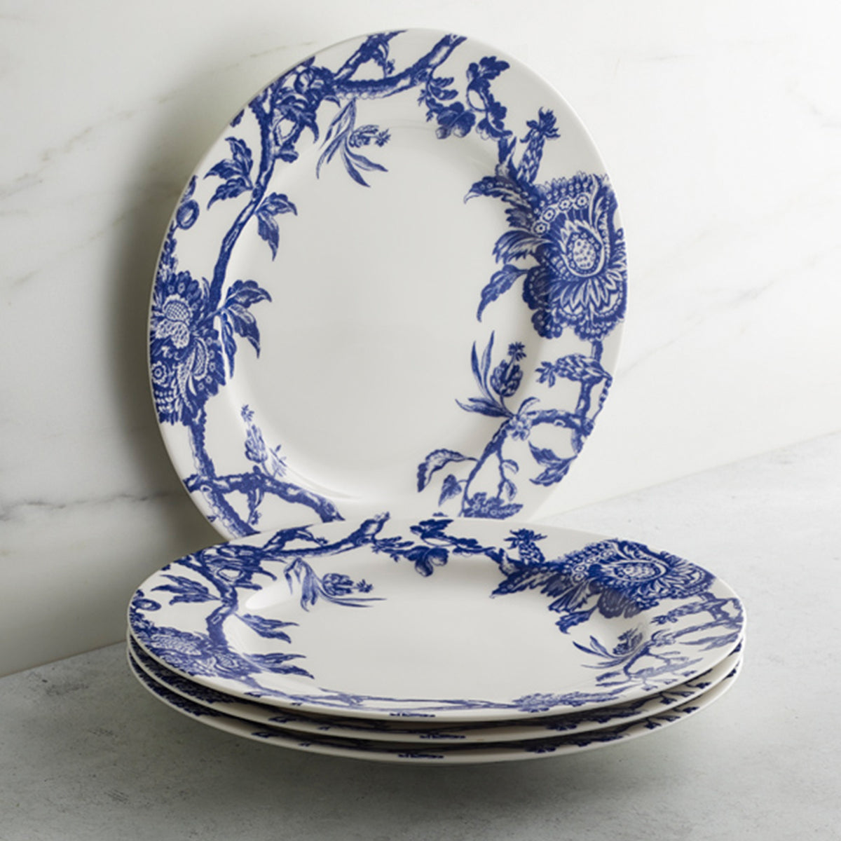 Four white plates with graphic blue floral patterns are stacked on a marble surface, showcasing Arcadia Rimmed Dinner Plate by Caskata Artisanal Home inspired by the Williamsburg Foundation.