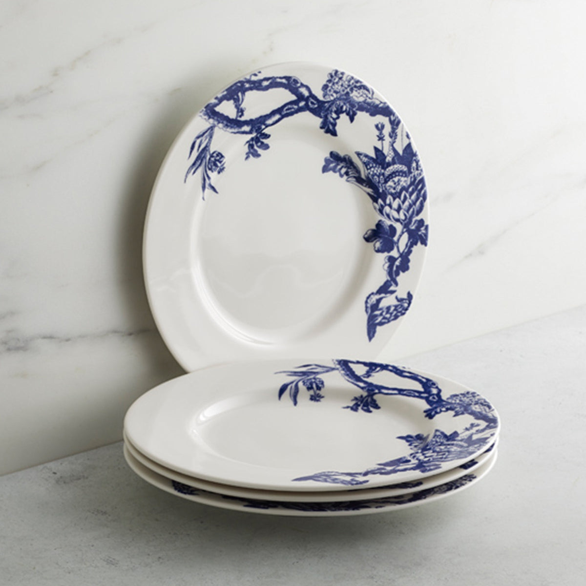 Three white ceramic Arcadia Rimmed Salad Plates by Caskata Artisanal Home, with intricate blue floral and bird designs, are stacked on a light grey surface.