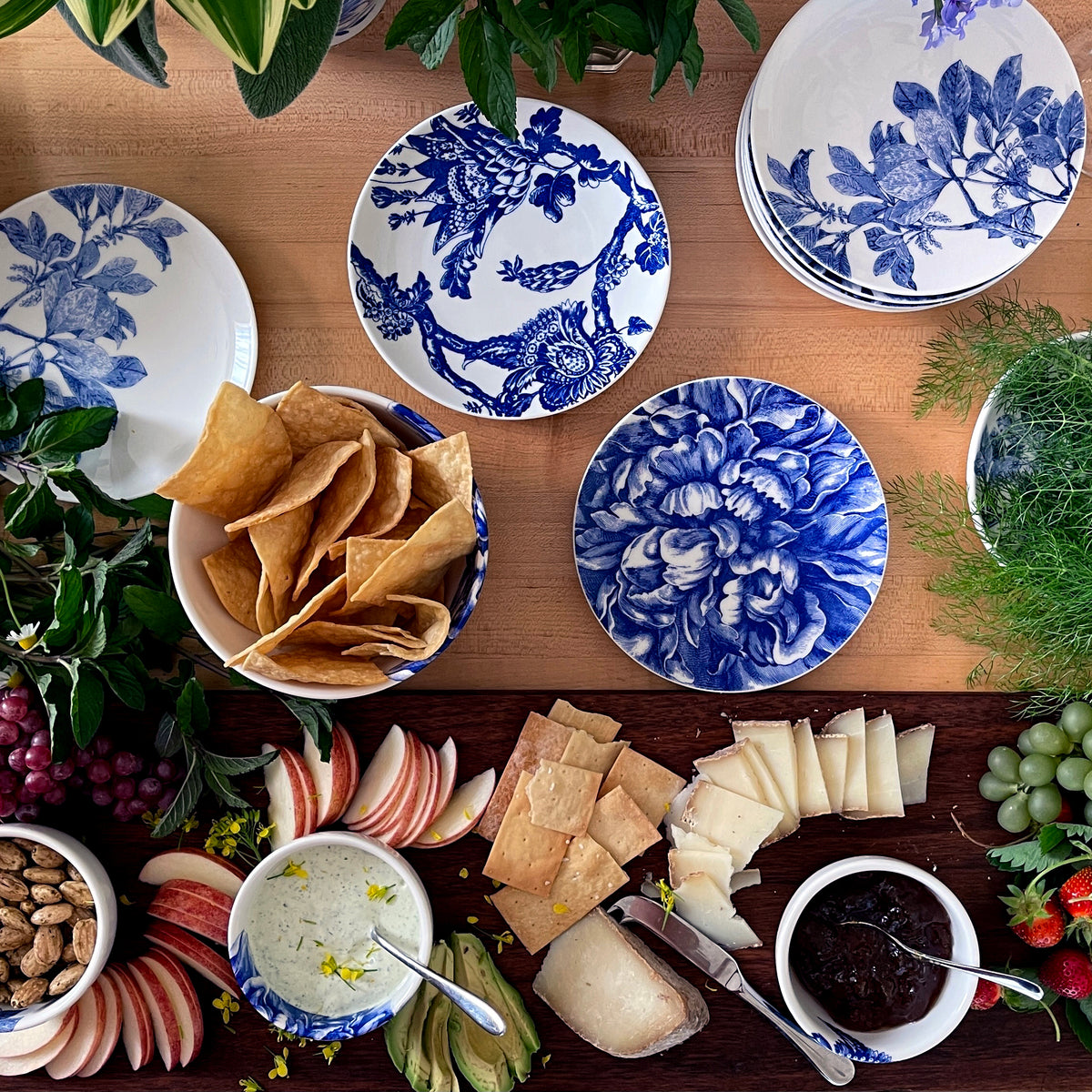 An overhead view of a wooden table showcases premium porcelain plates adorned with blue and white botanical details. The spread includes chips, cheese, apple slices, dip, and garnishes of fresh greens, grapes, and flowers— a perfect setting with Caskata Artisanal Home Arbor Blue Small Plates.