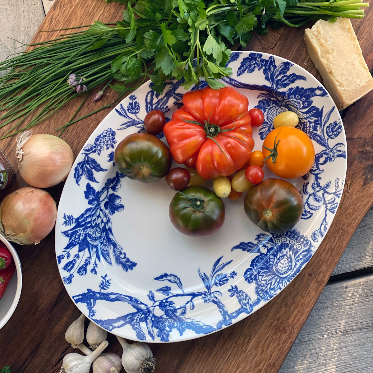 The Arcadia Large Oval Rimmed Platter by Caskata is displayed holding heirloom tomatoes on a cutting board surrounded by fresh vegetables.