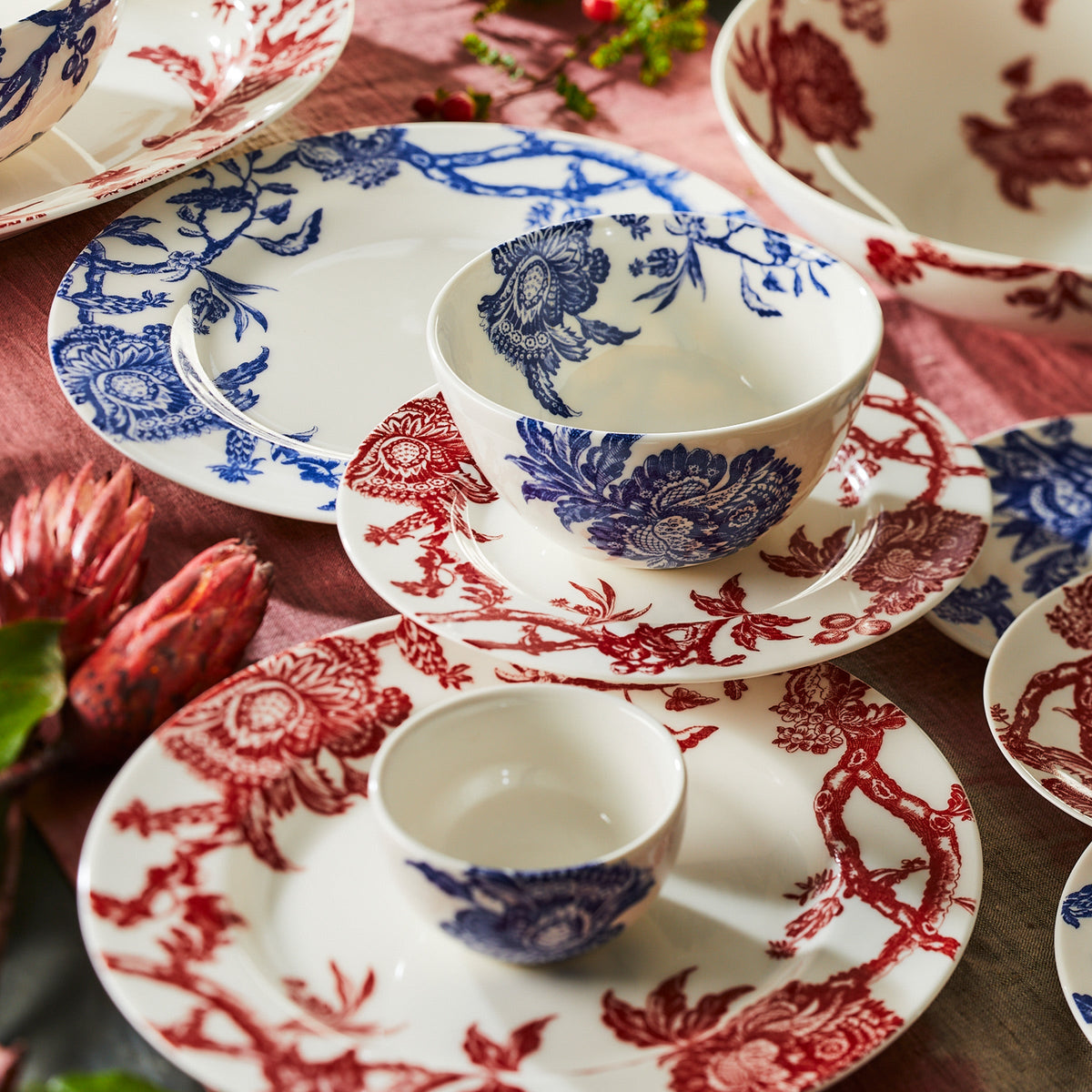 A table setting featuring premium porcelain dinnerware with white ceramic dishes adorned with intricate red and blue floral patterns. The set includes the Caskata Artisanal Home Arcadia Crimson Rimmed Salad Plates, bowls, and cups arranged on a pinkish tablecloth with floral accents, giving the display an heirloom feel.