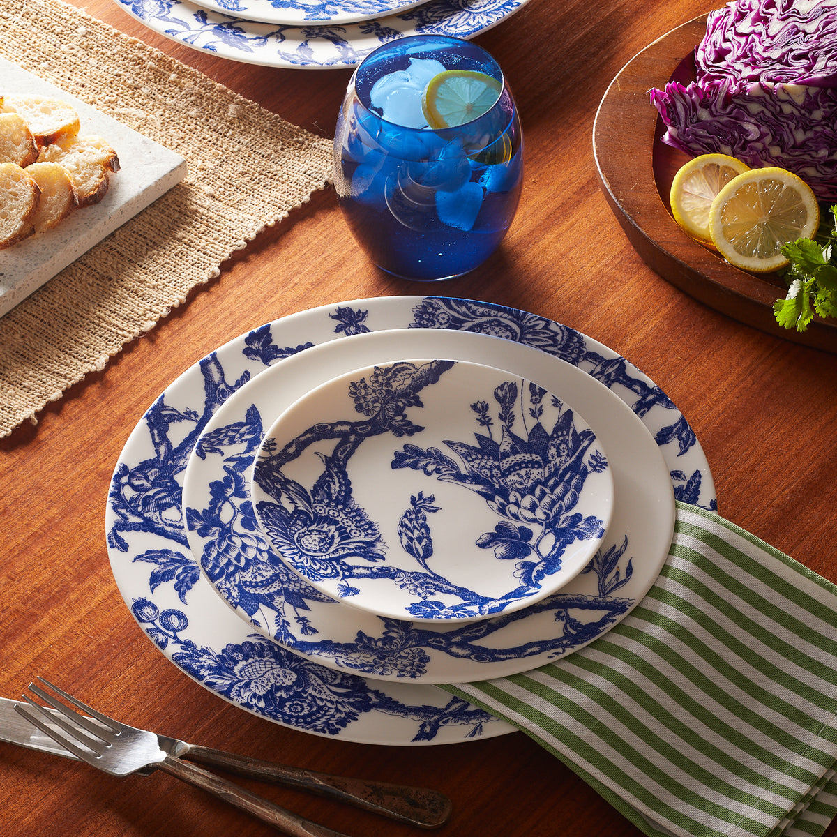 Table setting with Caskata Artisanal Home Arcadia Rimmed Dinner Plate in blue and white floral patterns, a green-striped napkin, a blue glass, sliced bread, and a bowl containing purple cabbage, lemon slices, and herbs on a wooden table.