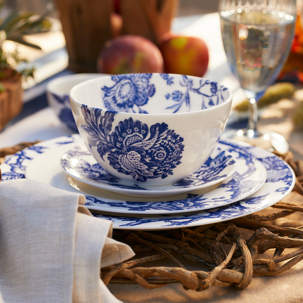 A white Arcadia Caskata porcelain dinnerware set with blue floral patterns, consisting of an Arcadia Cereal Bowl, a plate, and a saucer, placed on a table next to a folded napkin, a wine glass, and apples in the background.