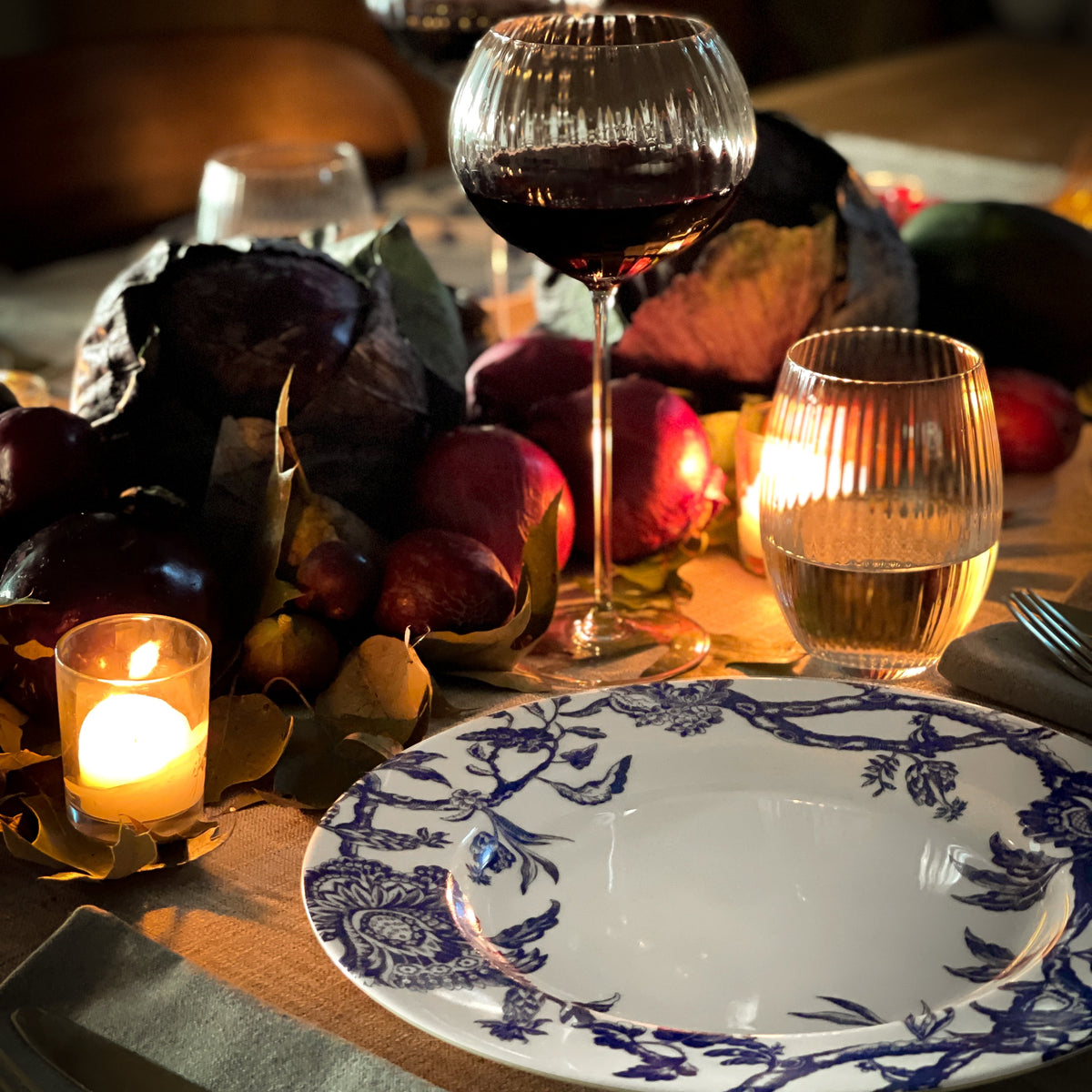 A set dining table features a blue and white Caskata Artisanal Home Arcadia Rimmed Dinner Plate, a glass of red wine, a glass of water, assorted vegetables, and a lit candle.