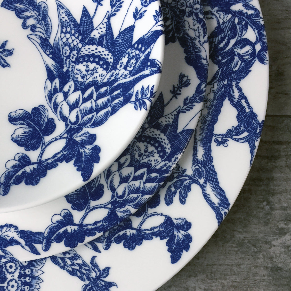 Close-up of three stacked Arcadia Small Plates by Caskata Artisanal Home with intricate blue floral patterns against a gray textured surface. Dishwasher and microwave safe, these heirloom-quality pieces add elegance to any table setting.