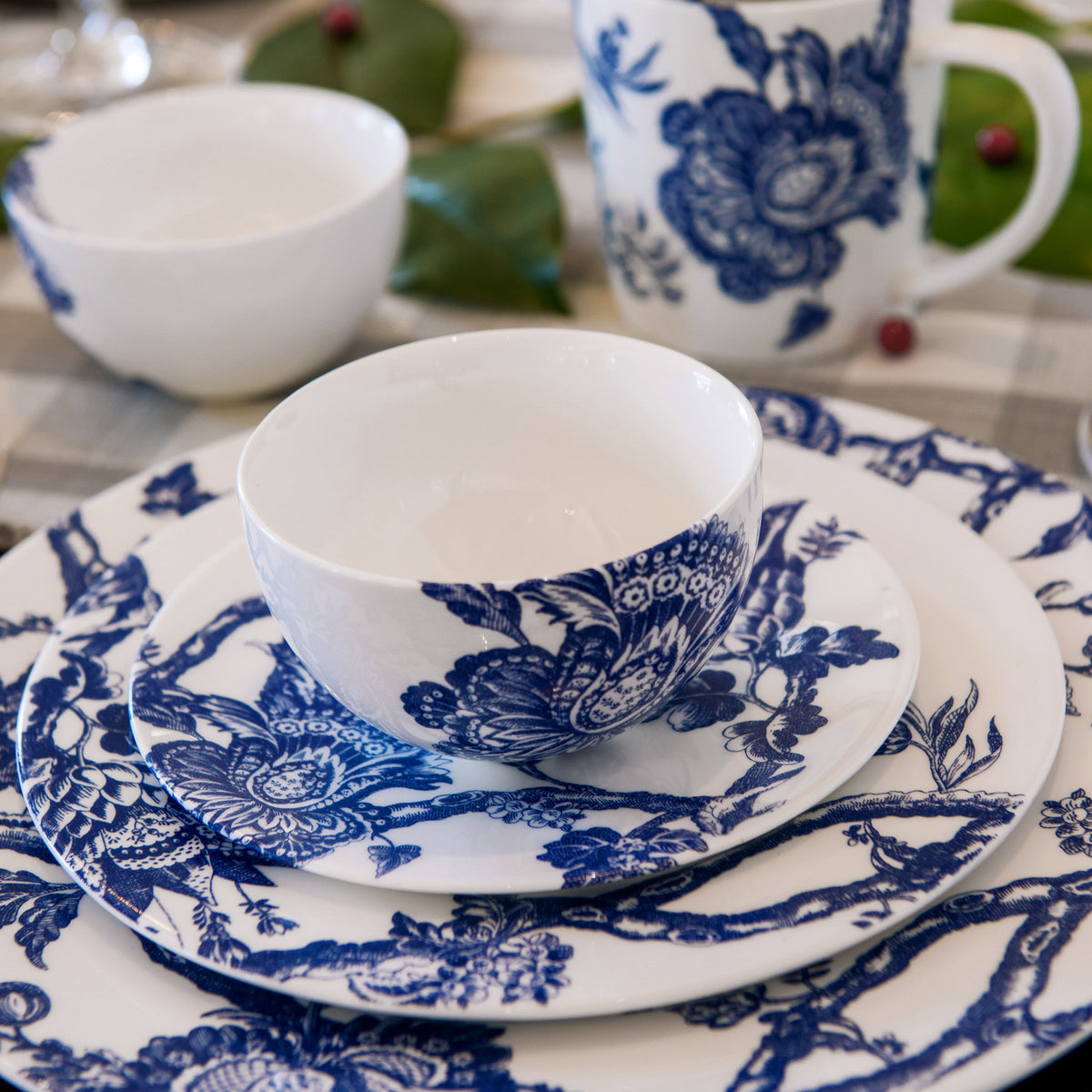 A set of Caskata Artisanal Home Arcadia Rimmed Salad Plates featuring intricate blue floral patterns, including plates, bowls, and a mug, beautifully arranged on a table with a checkered tablecloth.