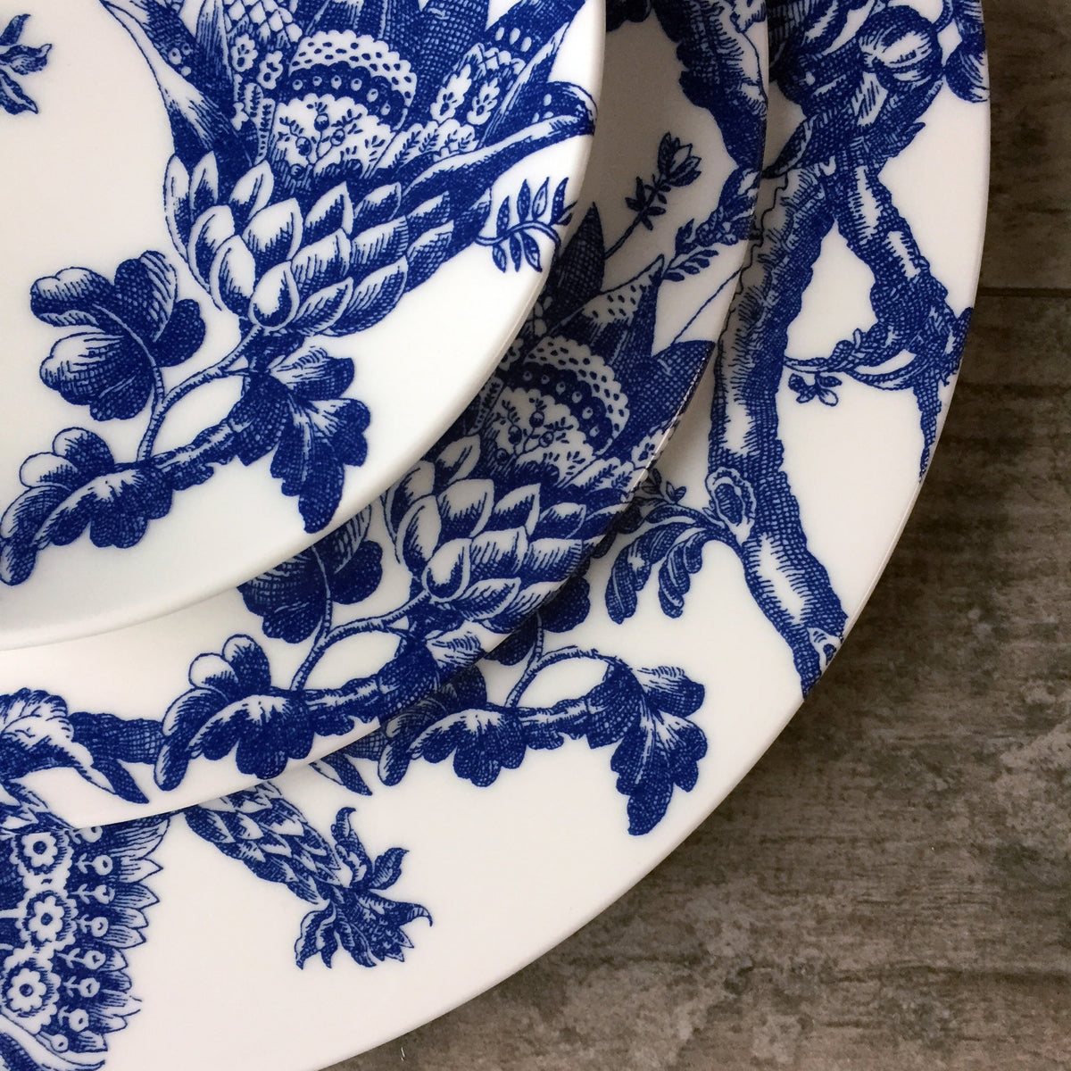 Close-up of three white ceramic plates with intricate blue graphic floral patterns, part of the Arcadia Rimmed Dinner Plate collection by Caskata Artisanal Home, stacked on top of each other on a wooden surface.