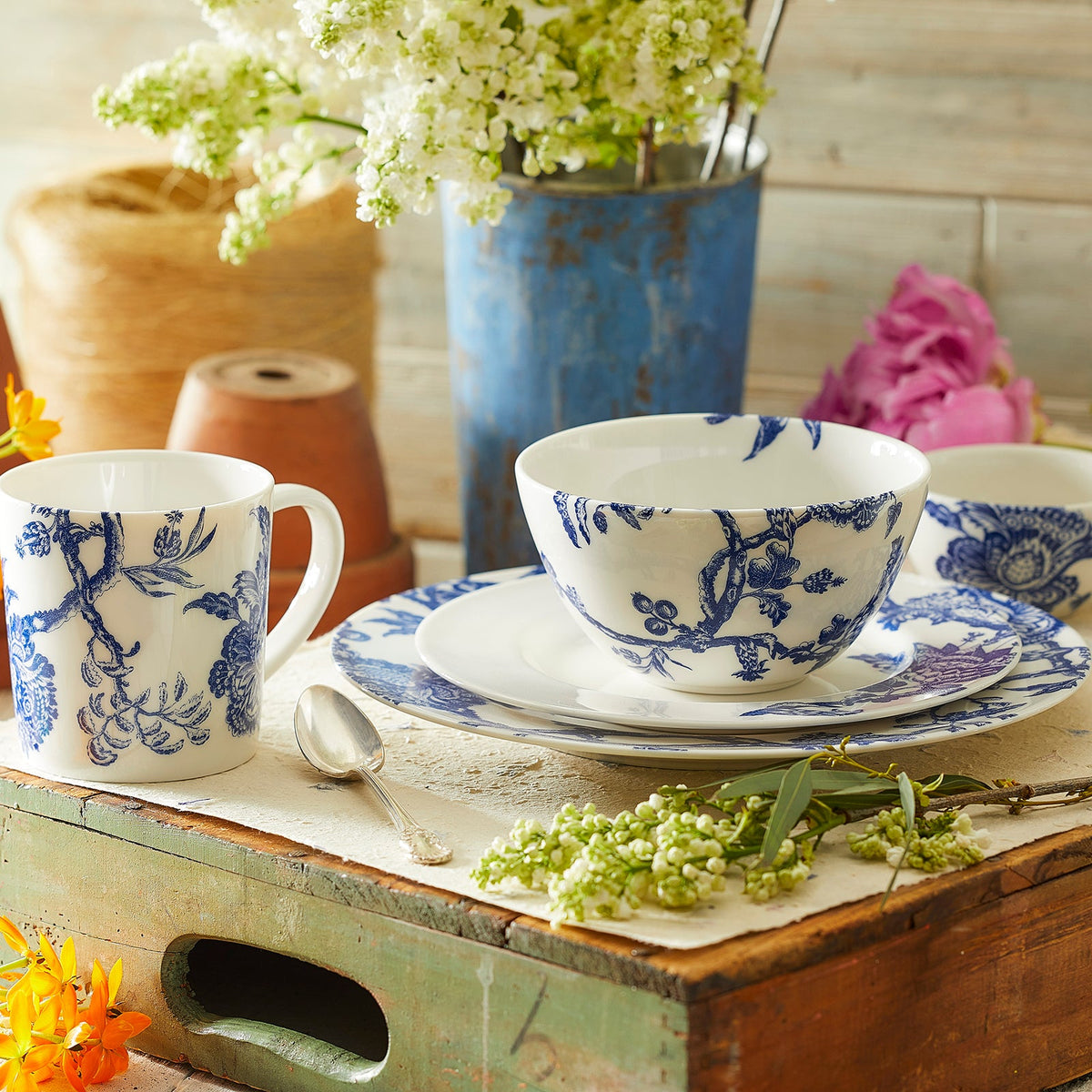 A rustic setting with blue and white Arcadia Rimmed Salad Plate from Caskata Artisanal Home, including a cup, bowl, and plates on a worn wooden box, surrounded by various flowers and a tin vase.