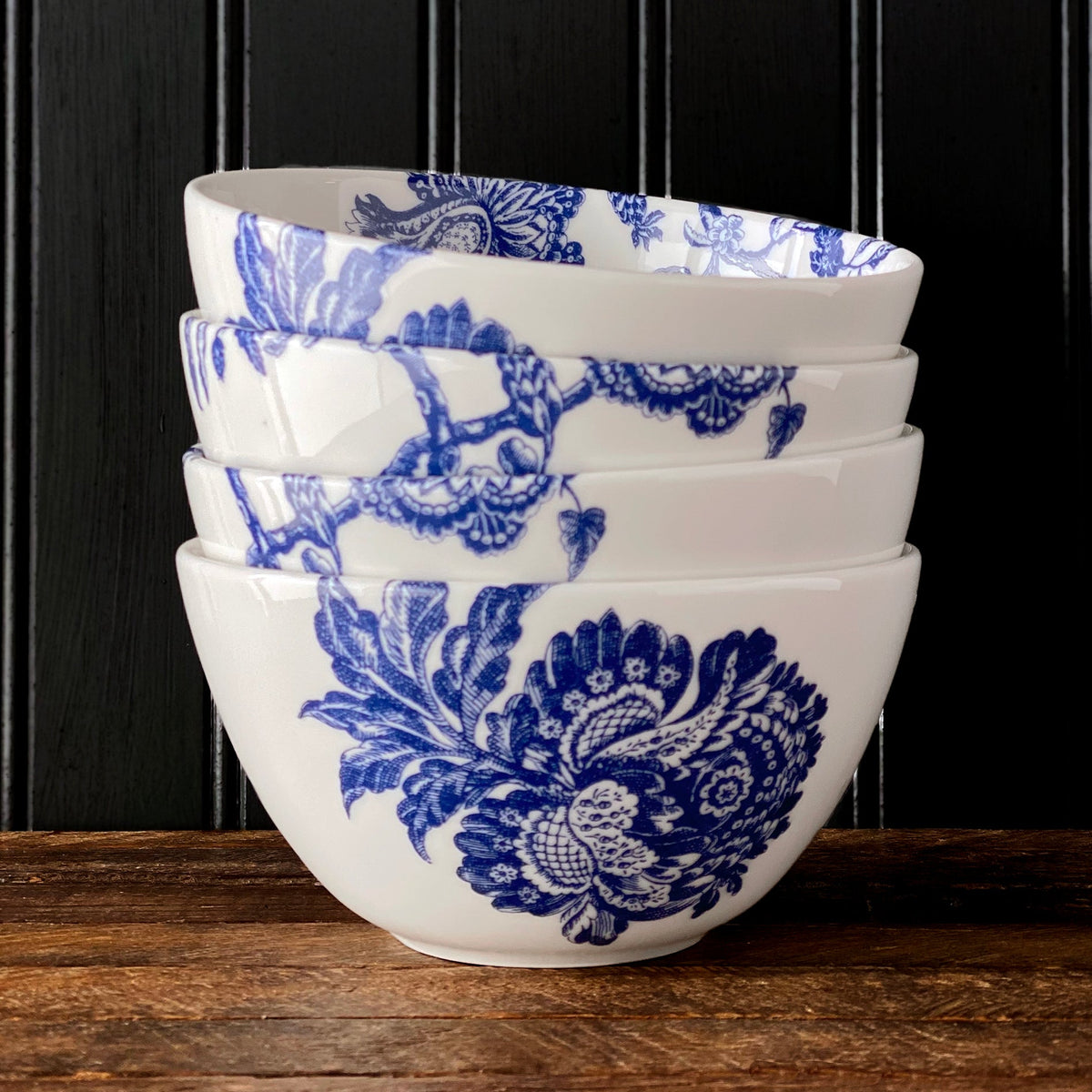 A stack of four white Arcadia Cereal Bowls by Caskata with blue floral patterns, part of the elegant Arcadia porcelain dinnerware collection, placed on a wooden surface against a black paneled background.