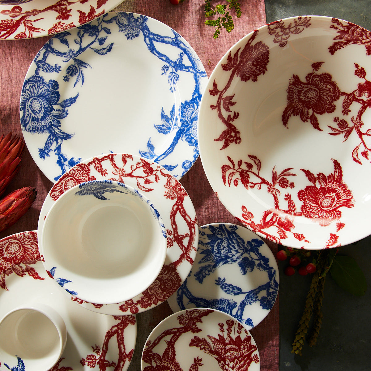 A collection of Arcadia Rimmed Dinner Plates by Caskata Artisanal Home with intricate blue and red graphic florals, arranged on a maroon tablecloth.