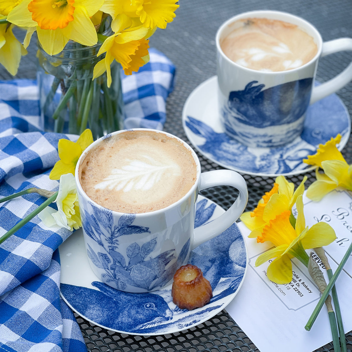 Two cups of cappuccino with latte art in blue floral mugs on matching saucers, set on a table adorned with yellow daffodils, a blue checkered cloth, and a small pastry atop **Bunnies Small Plates by Caskata Artisanal Home**.