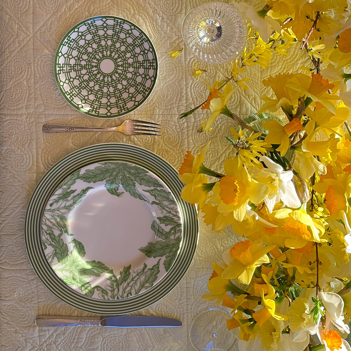 A table setting with green and white patterned high-fired Caskata Artisanal Home Newport Stripe Verde Rimmed Dinner Plates, silverware, and a glass placed next to a bouquet of yellow flowers on a yellow quilted tablecloth.
