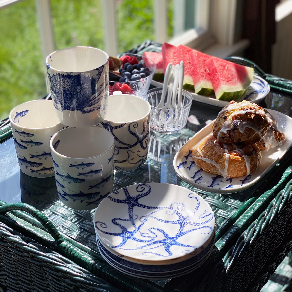 A table set with heirloom-quality dishware featuring marine life designs, croissants and pastries on a plate, fresh berries in bowls, **Caskata Artisanal Home Starfish Small Plates** alongside watermelon slices on a tray, and plastic forks in a glass.