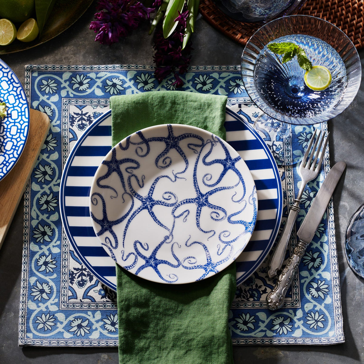 A dinner setting with Caskata Artisanal Home&#39;s premium porcelain Starfish Coupe Salad Plate featuring starfish and striped designs, a green napkin, silverware, and a cocktail with lime and mint, arranged on a decorative placemat.