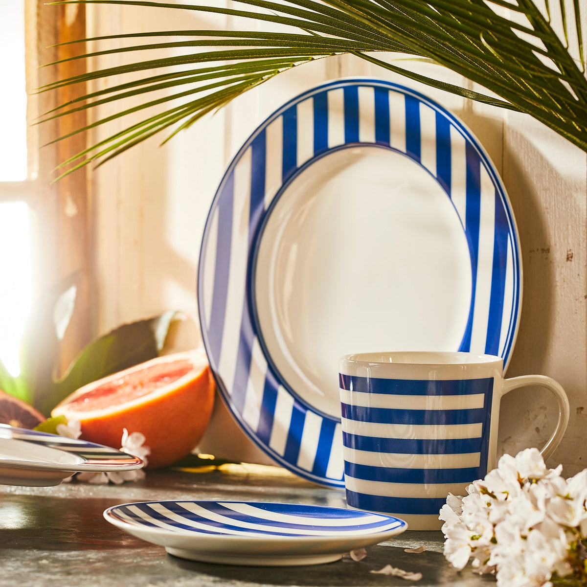 A blue and white striped dinnerware set, including a plate, a bowl, and a cup, arranged on a table near a sliced grapefruit and flowers with sunlight streaming through a window. The Beach Towel Stripe Small Plates by Caskata Artisanal Home add an elegant touch to the sunny breakfast scene.