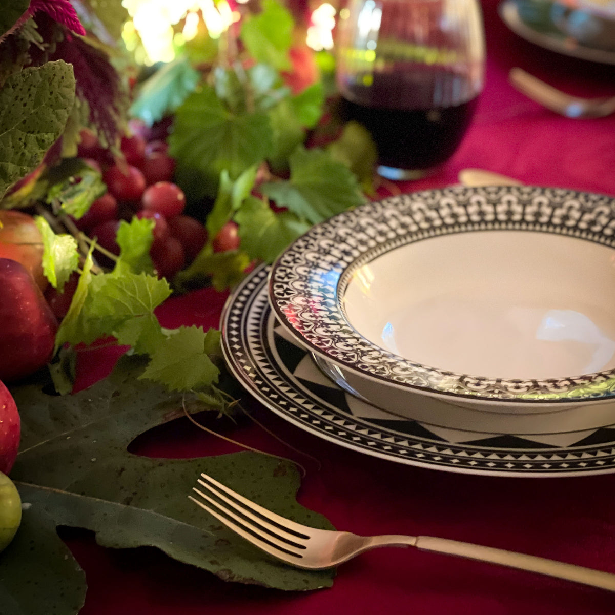 Elegant table setting with sophisticated dinnerware, a Fez Rimmed Dinner Plate by Caskata Artisanal Home featuring Moroccan patterns, a fork, glass of red wine, and an arrangement of fruits and greenery on a red tablecloth.
