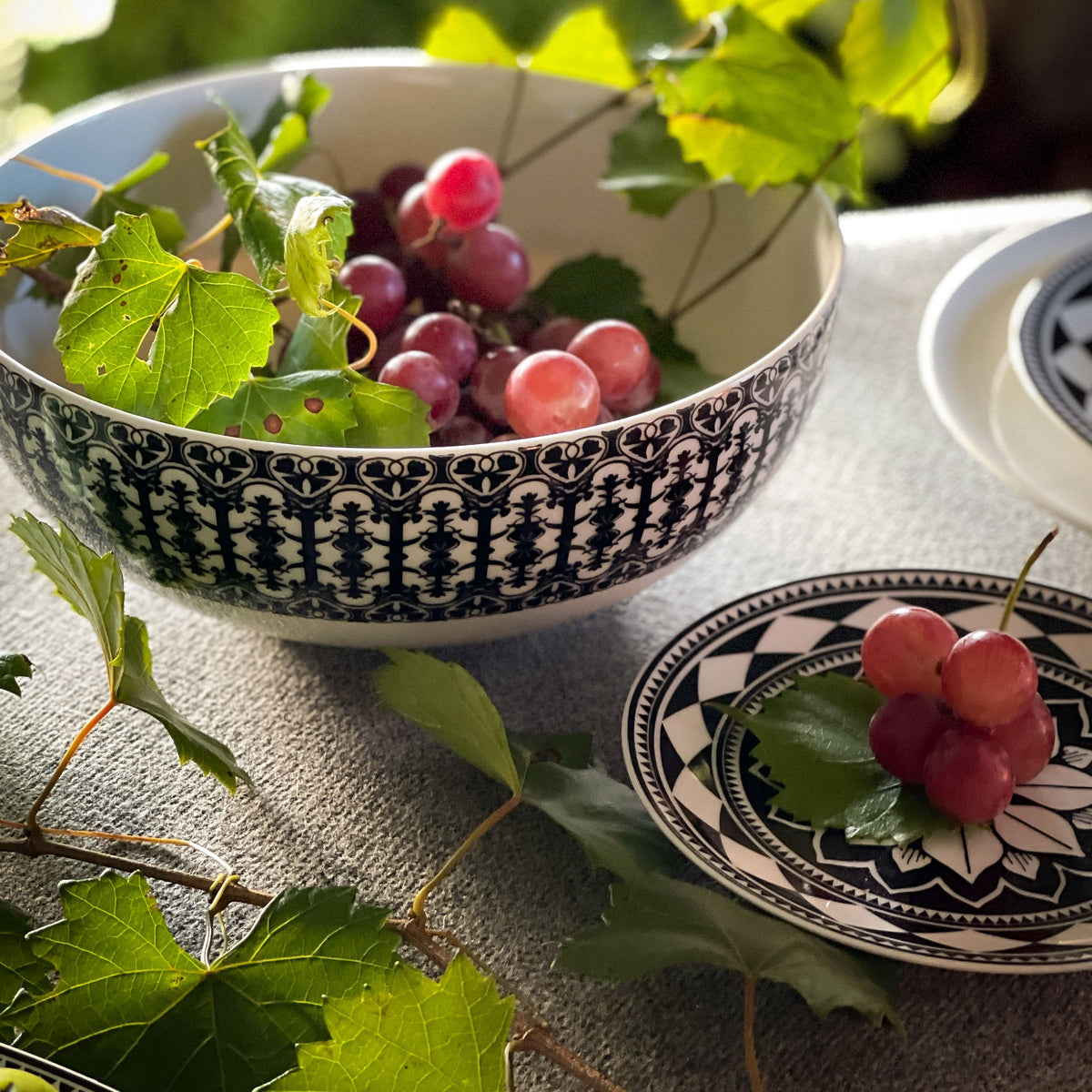 A patterned bowl and Fez Small Plates by Caskata Artisanal Home, showcasing bold geometrics, hold red grapes and grape leaves on a tablecloth, with sunlight providing illumination.