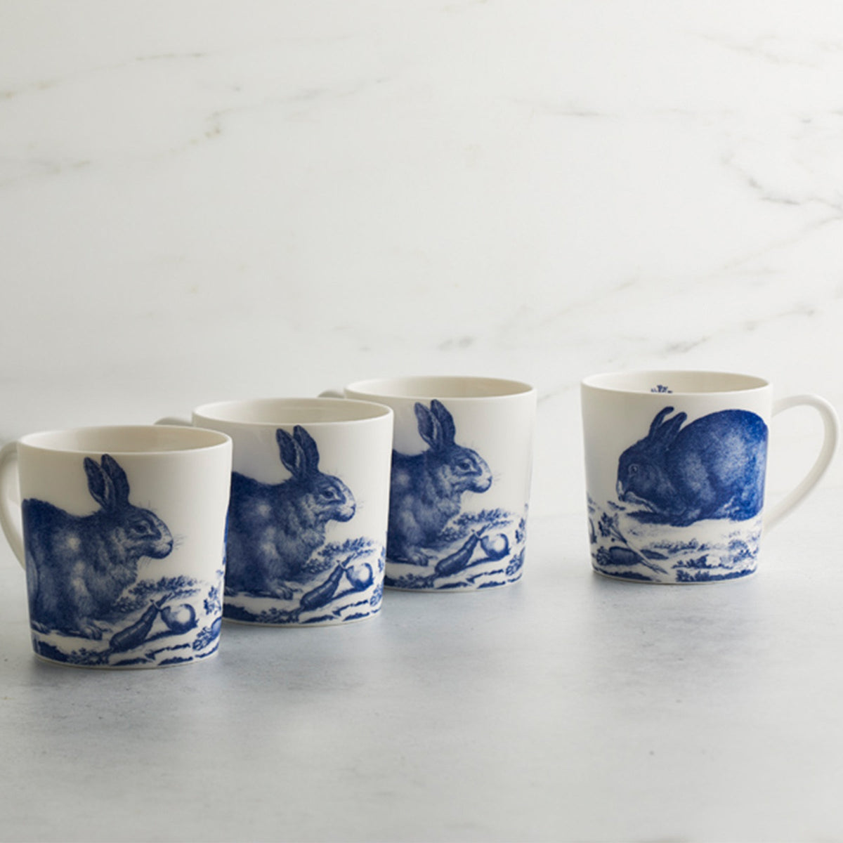 Four white high-fired porcelain Bunnies Mugs with blue illustrations of rabbits standing in a row on a light surface against a marble background, evoking a charming, Spring-inspired aesthetic from Caskata Artisanal Home.