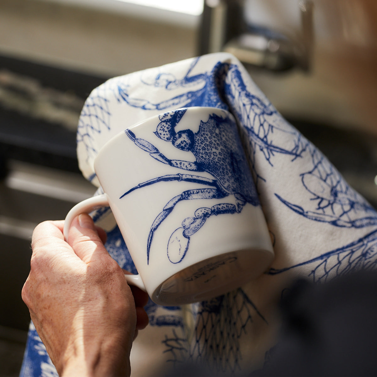 A person is drying a high-fired porcelain Crab Mug from Caskata Artisanal Home with a dish towel featuring similar blue designs.