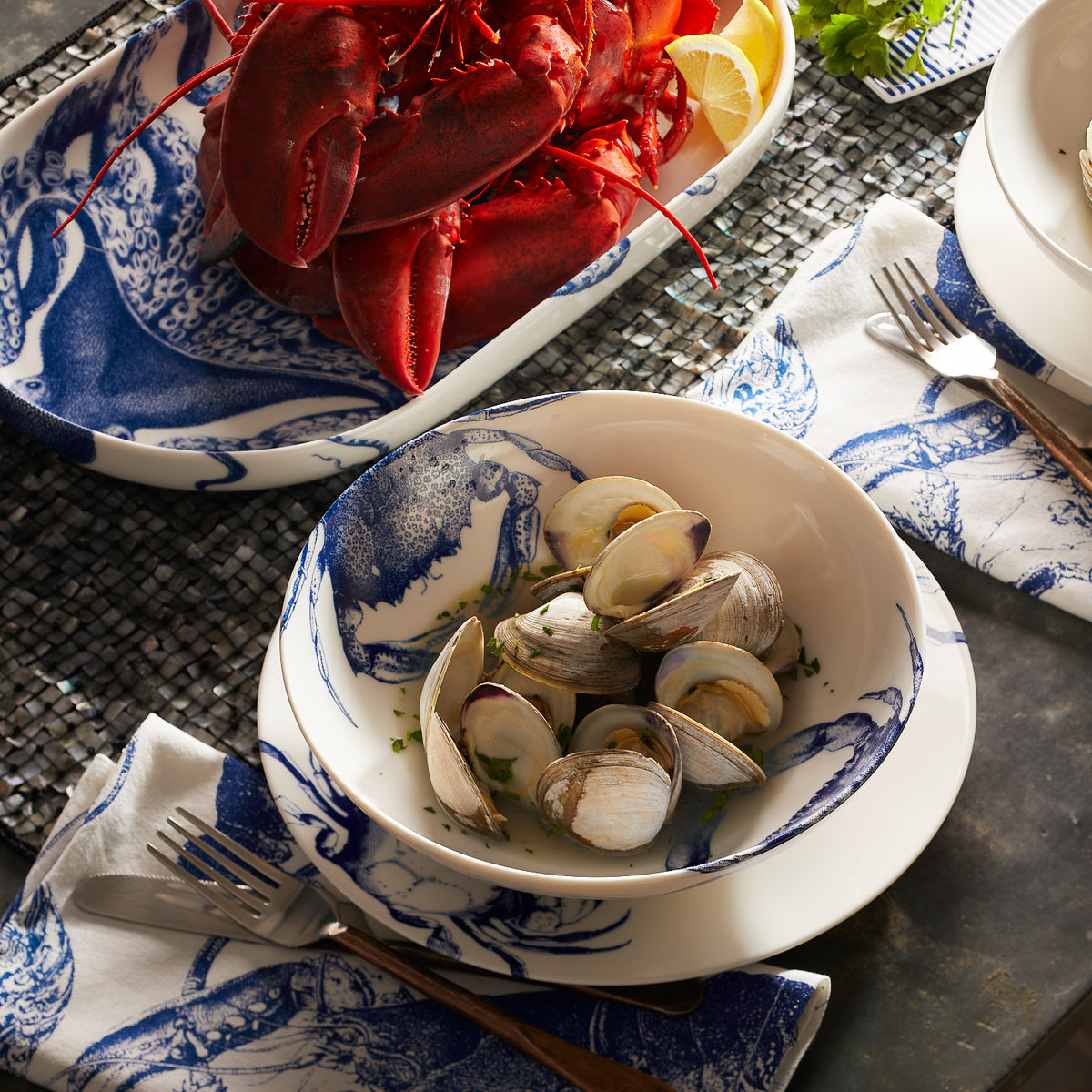 A bowl of clams in a blue and white high-fired porcelain Crab Entrée Bowl with a crab pattern from Caskata Artisanal Home, next to a platter of cooked lobsters with lemon slices, is placed on a table with silverware and napkins. The set is both dishwasher and microwave safe.