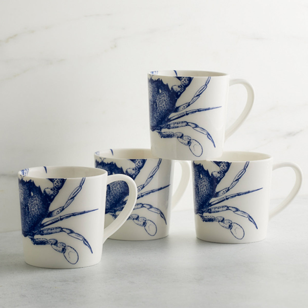 Four white, high-fired porcelain mugs with blue crab illustrations are stacked in a pyramid formation on a marble surface. These Caskata Artisanal Home Crab Mugs are not only visually appealing but also dishwasher safe for easy cleaning.