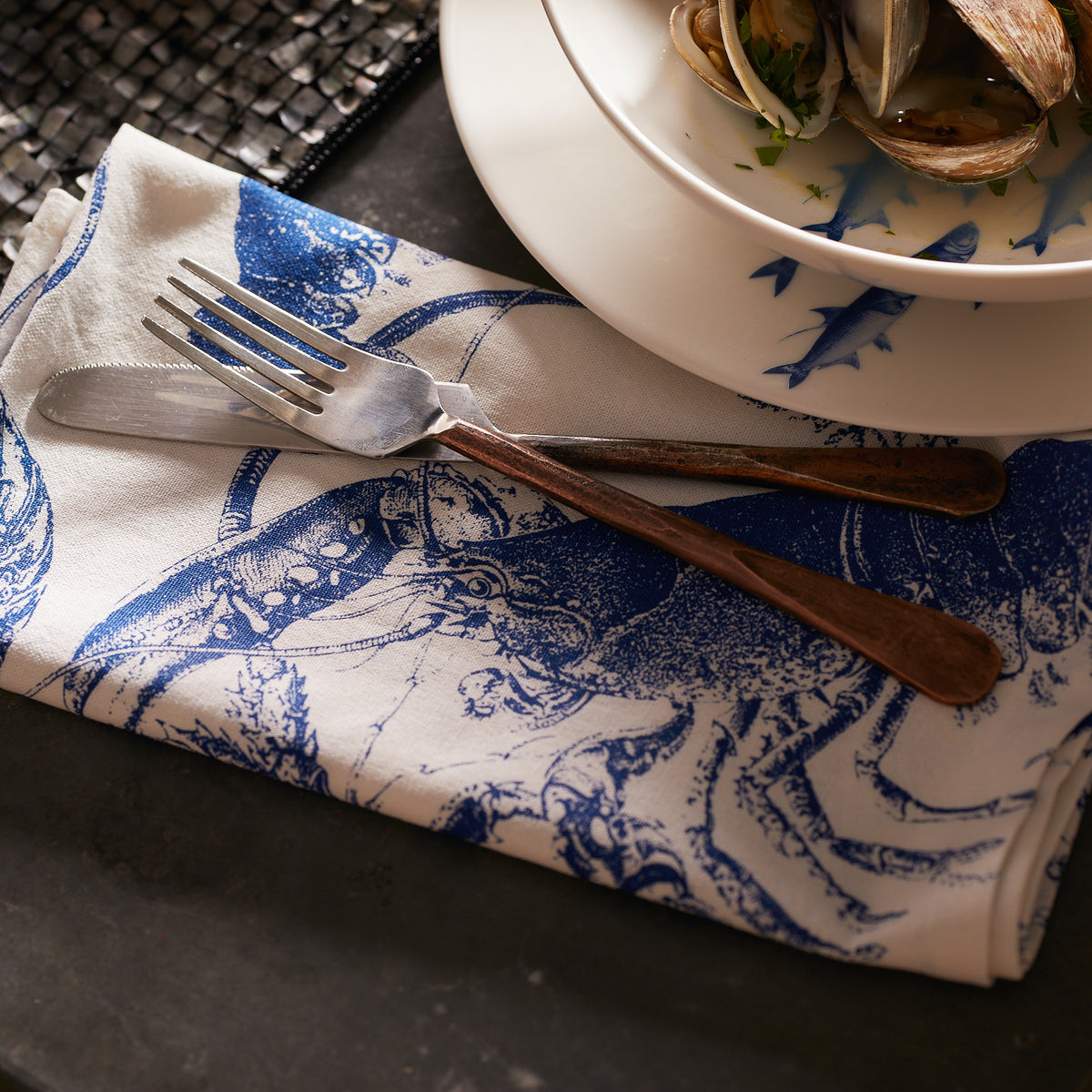 A Blue and White 100% cotton lobster dinner napkin from Caskata folded under a knife and fork.