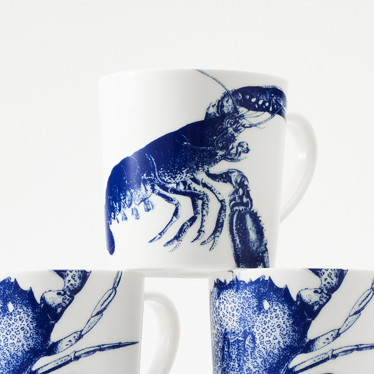 Three high-fired porcelain **Lobster Mugs** featuring a blue lobster design from **Caskata Artisanal Home** are stacked, with the top mug prominently displaying the full lobster image. Made in Sri Lanka, these unique lobster mugs add a touch of coastal charm to any kitchen.
