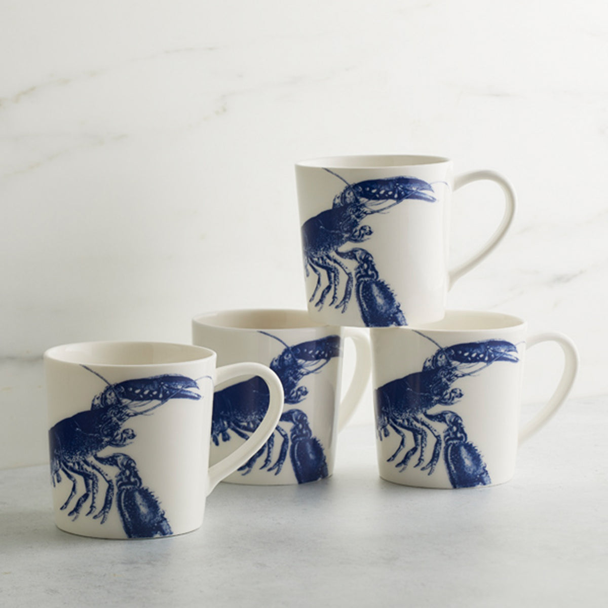 Four white high-fired porcelain Lobster Mugs from Caskata Artisanal Home with blue illustrations are stacked in a pyramid on a light-colored surface. Made in Sri Lanka, these elegant pieces add a coastal touch to any kitchen.