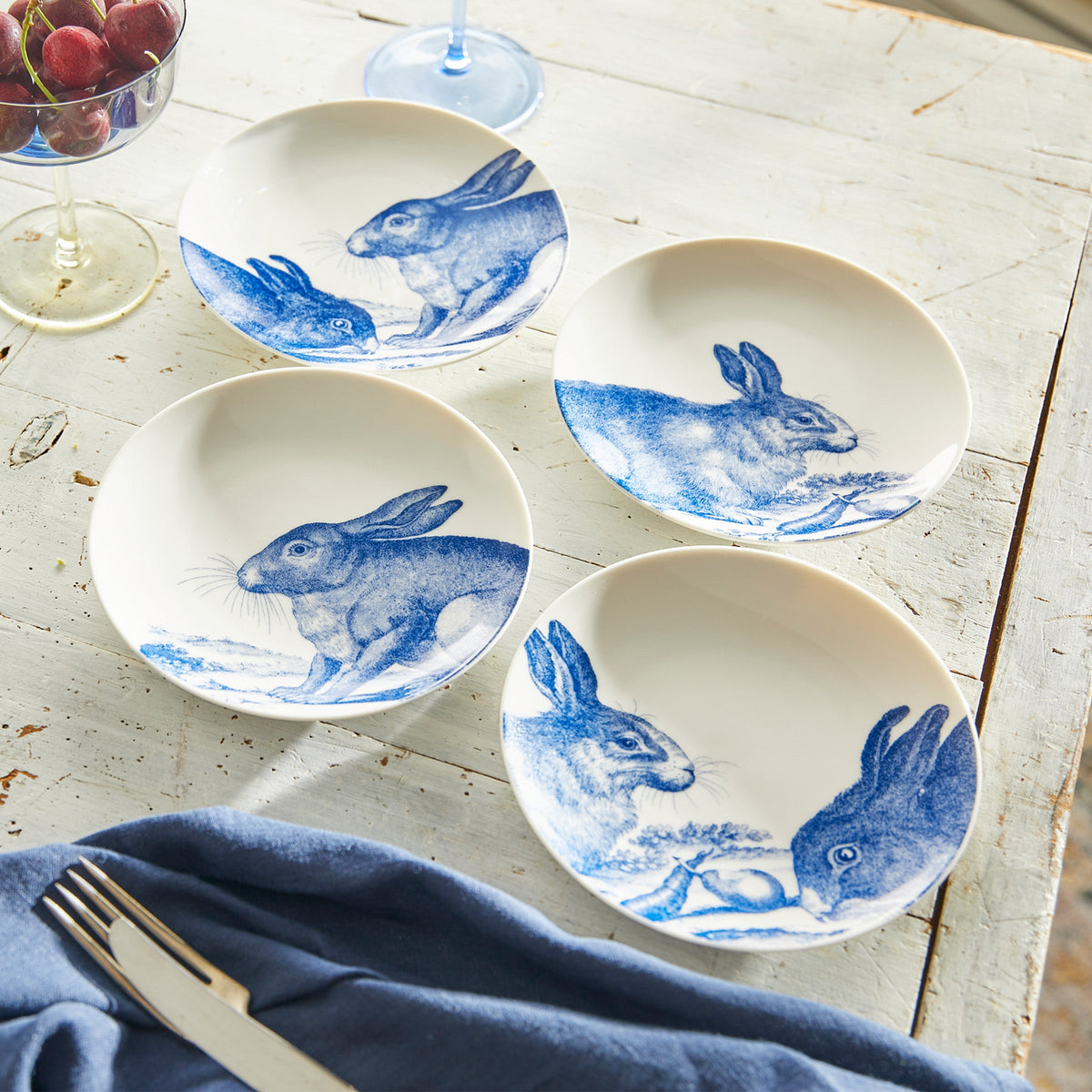 Four Bunnies Small Plates made of high-fired porcelain with blue drawings of rabbits from Caskata Artisanal Home are arranged on a distressed white wooden table. A glass of red grapes, a fork, a knife, and a blue cloth are also on the table.