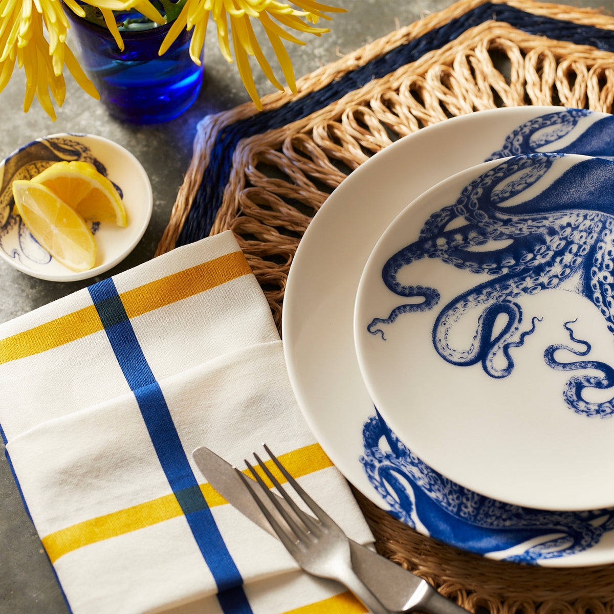 A table setting with blue and white Lucy Coupe Dinner Plates by Caskata Artisanal Home, a yellow and blue checked napkin, a fork and knife, a small dish with lemon wedges, and a blue vase with yellow flowers.