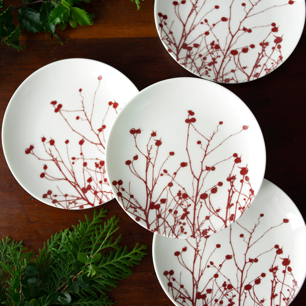 Four white porcelain Winterberries Small Plates by Caskata Artisanal Home, with red botanical designs reminiscent of Winter berries, are arranged on a wooden table with some green foliage.