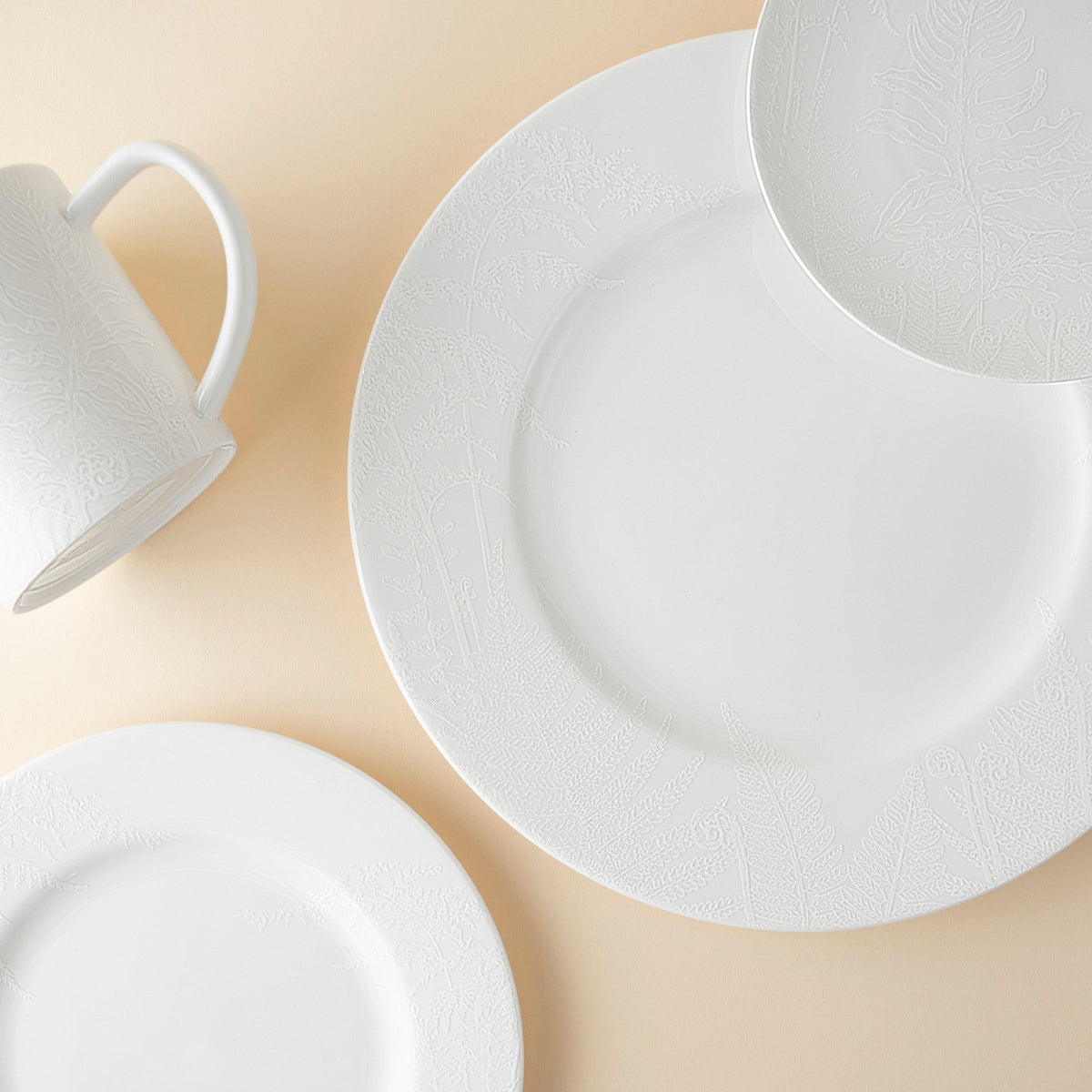 Three **Spring Small Plates by Caskata Artisanal Home** and a white mug with delicate embossed fern patterns are arranged on a light beige background, showcasing the elegance of botanical imagery in our Spring collection.