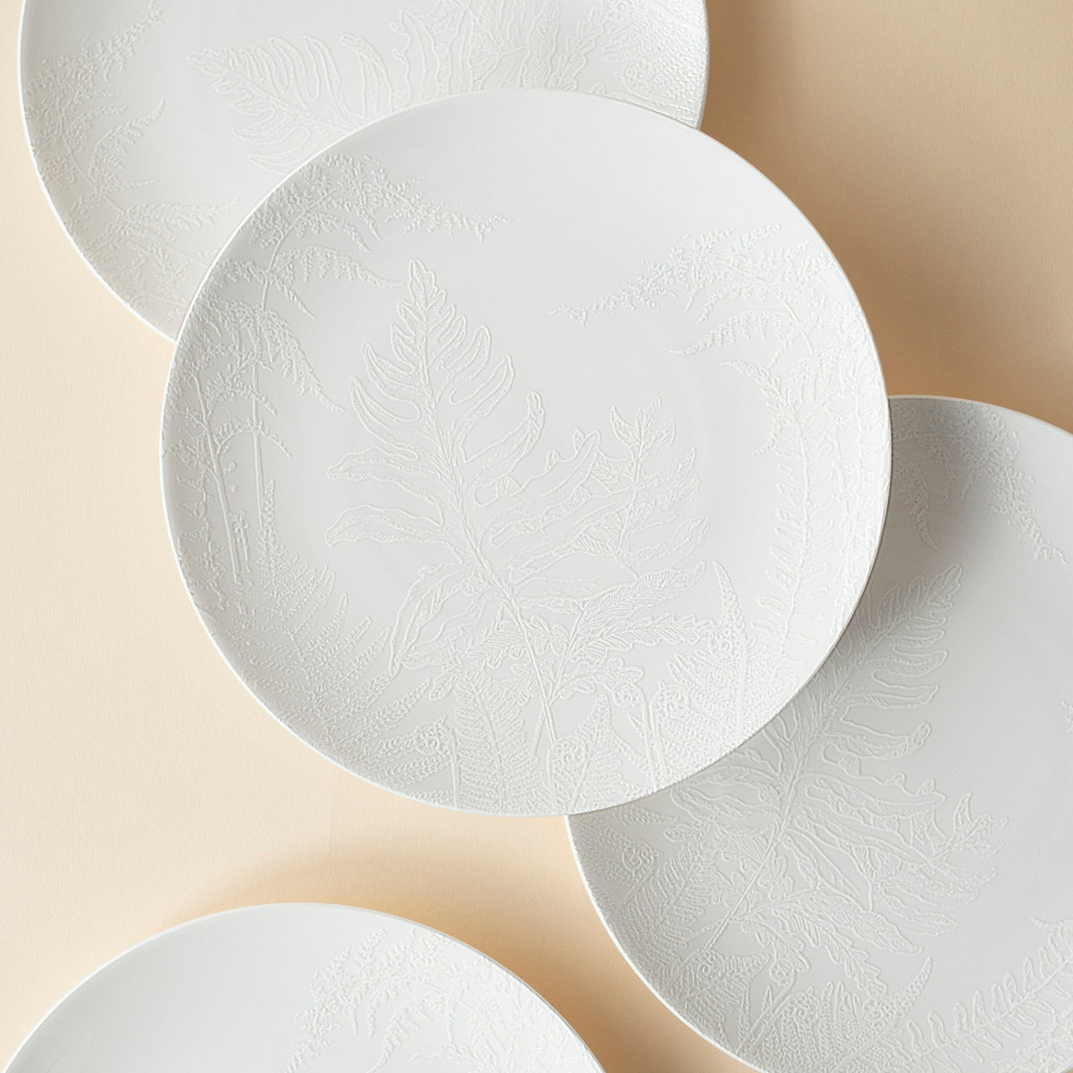 Four white porcelain plates with subtle, textured botanical patterns are displayed overlapping on a beige surface, perfect additions to your Spring collection. These are the exquisite *Spring Small Plates* from *Caskata Artisanal Home*.