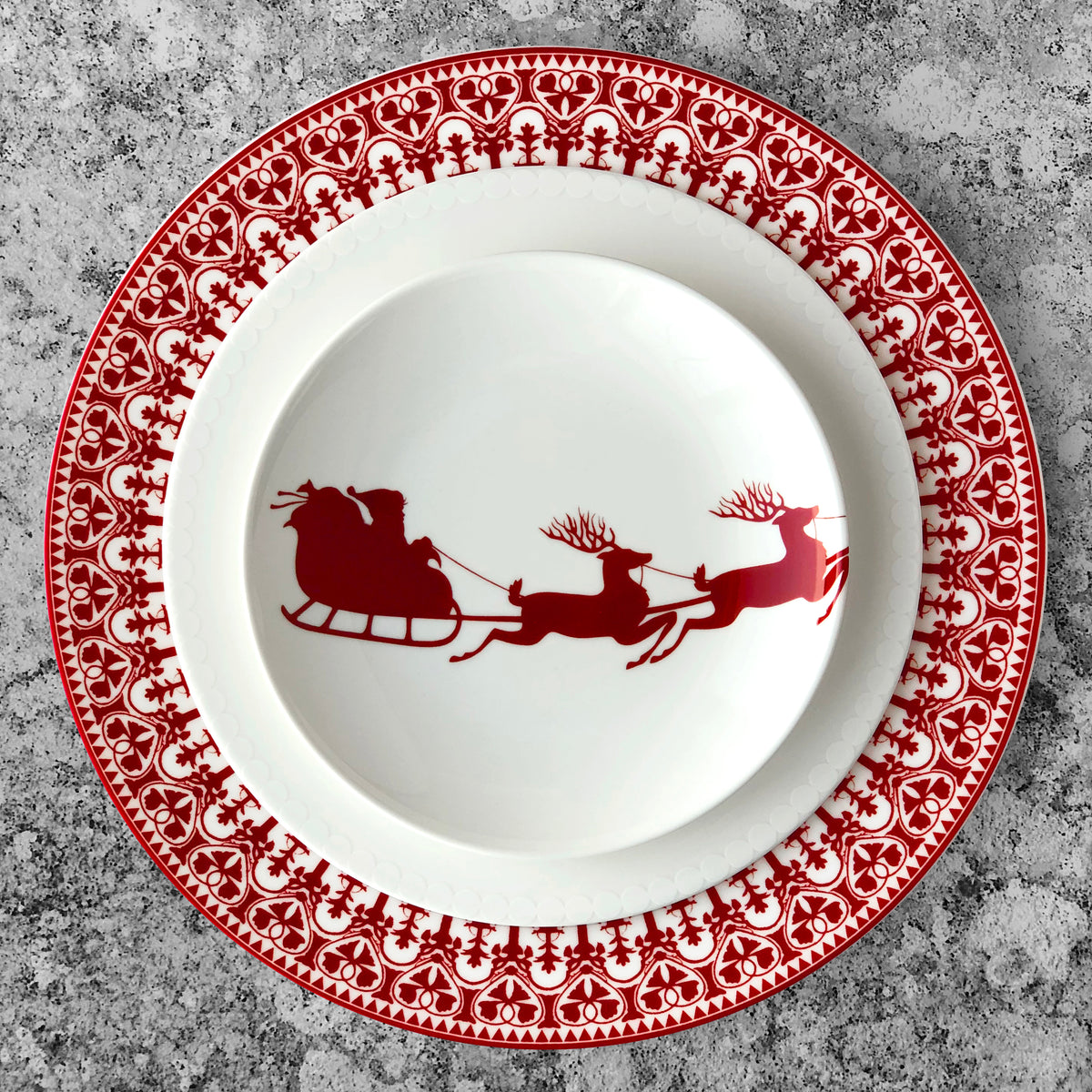 A white plate with red rim features a silhouette of Santa&#39;s sleigh pulled by two reindeer. The Sleigh Small Plates, part of heirloom-quality dinnerware by Caskata Artisanal Home, sit on a larger red and white decorative plate with a snowflake pattern on a gray background. These Christmas canapé plates come in boxed sets of 4.