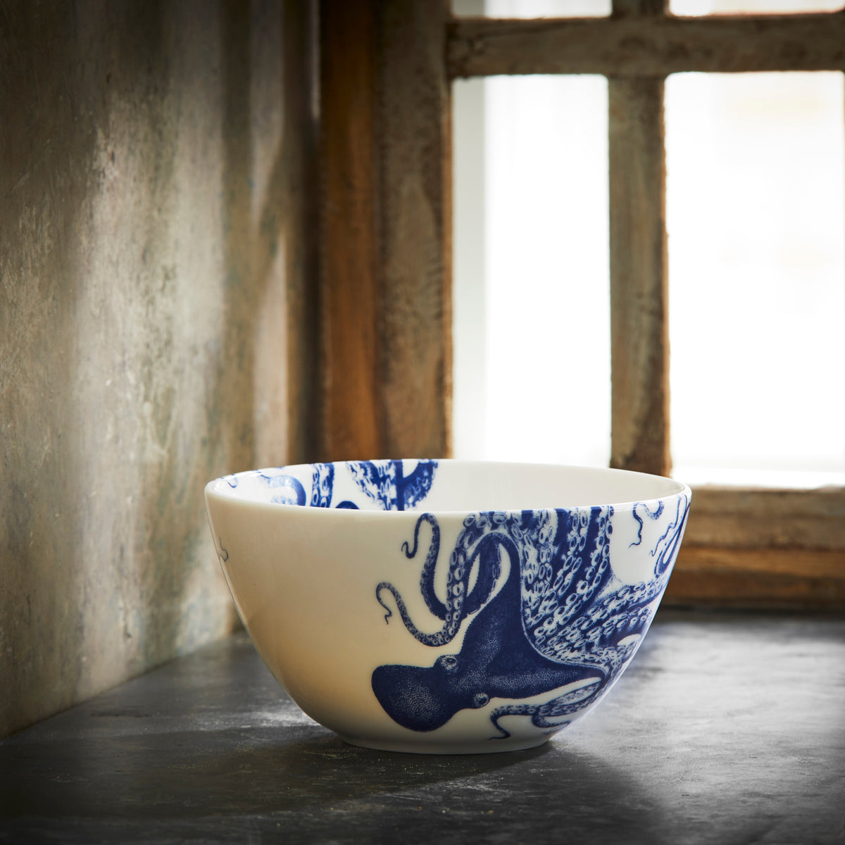 A Caskata Lucy Cereal Bowl featuring Lucy the octopus is placed on a dark countertop near a window with natural light streaming through.